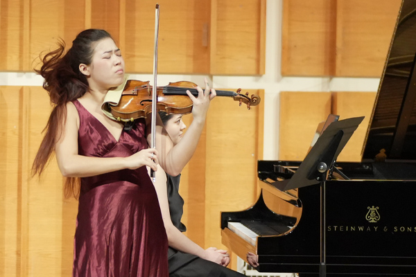 Young woman in a burgundy silk dress playing violin, her hair showing movement and a piano behind her