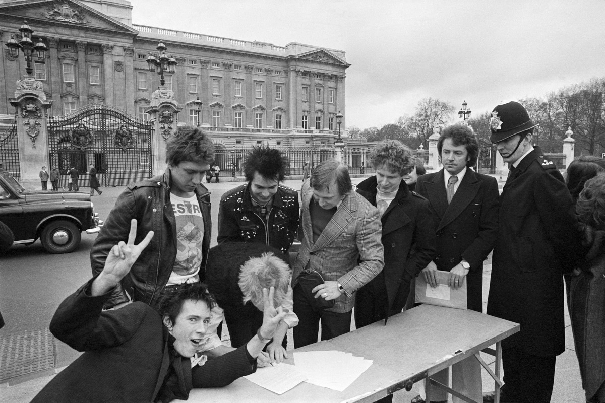 The Sex Pistols outside Buckingham Palace, signing a contract on a table with a policeman looking over them, 1977