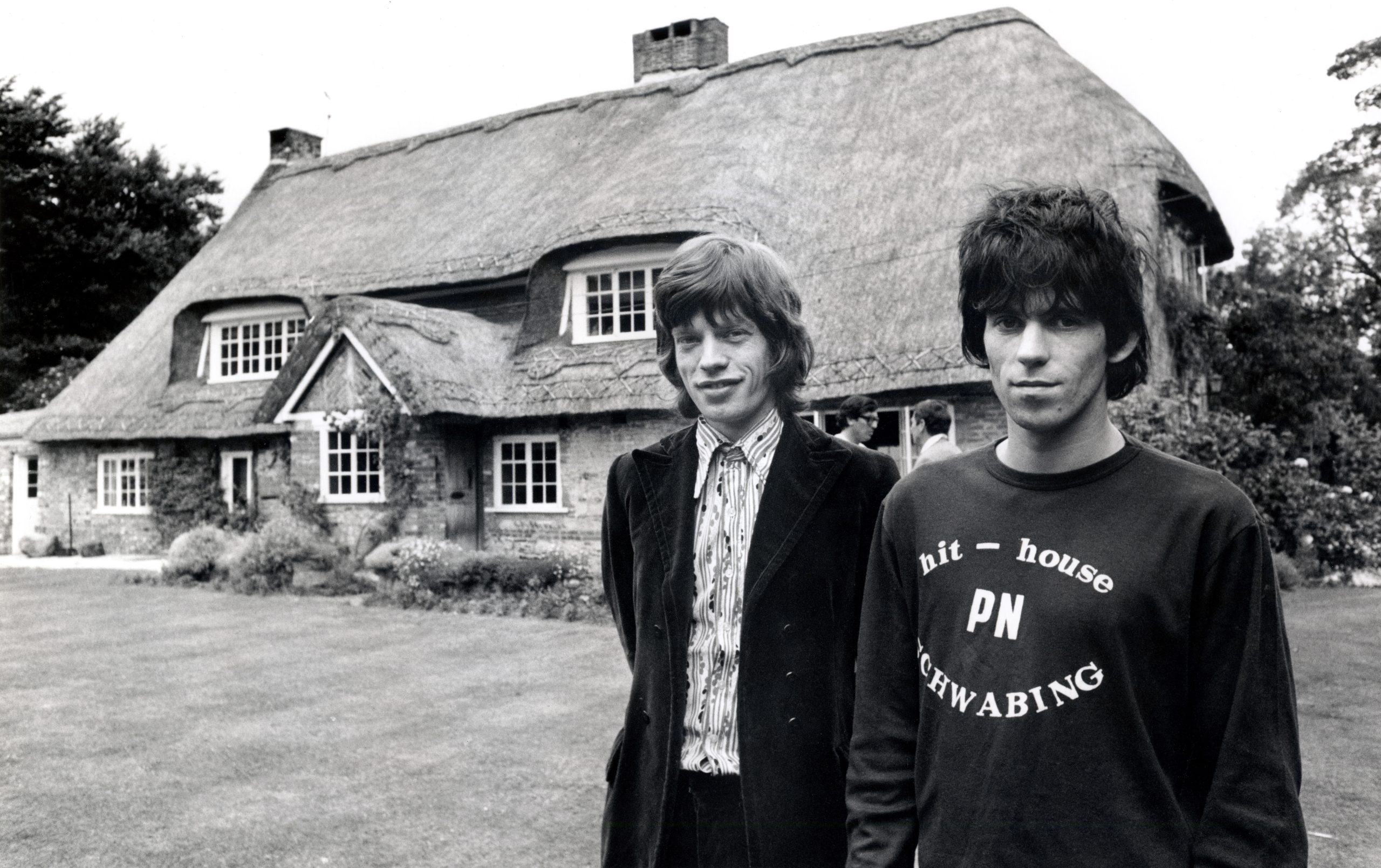 Black and white photo of Mick Jagger (left) and Keith Richards from The Rolling Stones stand in front of 'Redlands', Keith Richards' Sussex house, Mick in a suit and Keith in a sweatshirt
