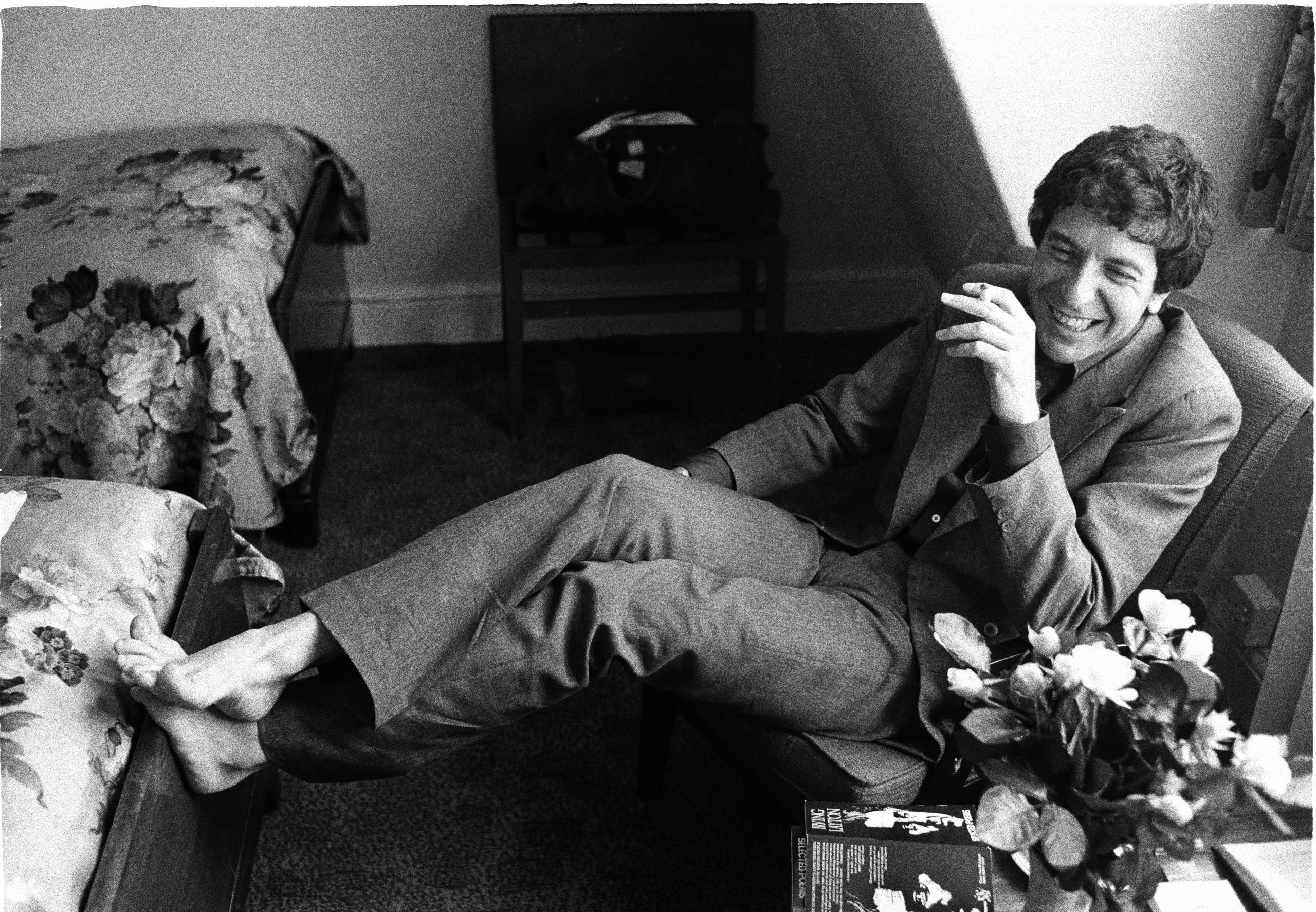 Black and white photo of Leonard Cohen in a hotel room with his feet on the bed smoking a cigarette