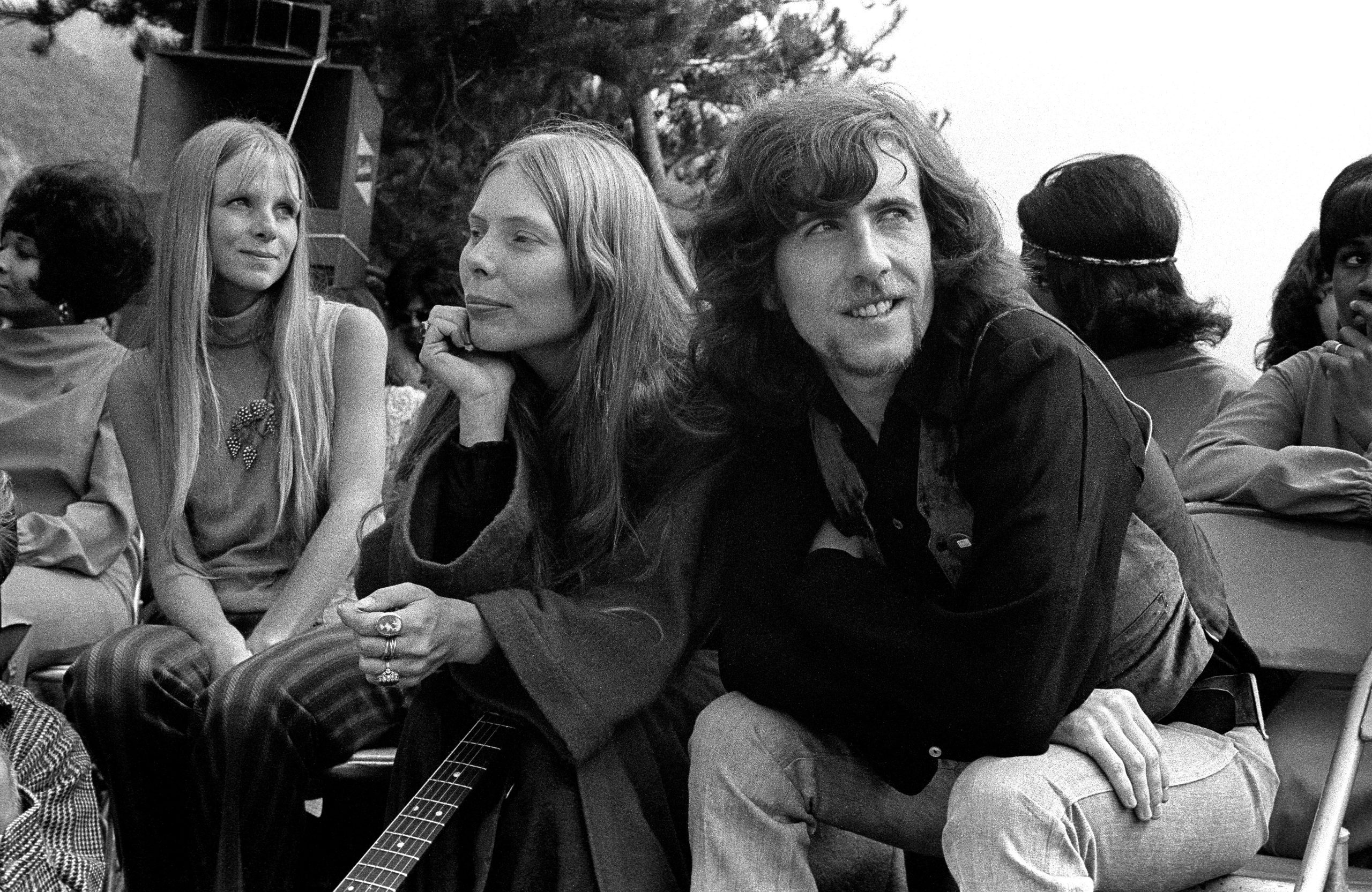 Black and white photo of Graham Nash and Joni Mitchell sitting backstage with a guitar and other people around